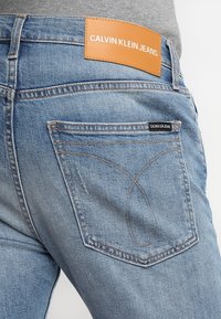 Blå denimjeans med hög midja, orange läderbakficka med "CALVIN KLEIN JEANS" och ett unikt sömnadsmönster på fickorna.