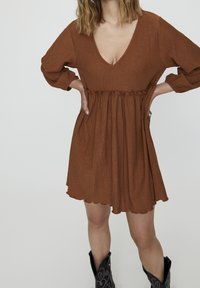 Rostfarbenes Kleid mit V-Ausschnitt, langen Ärmeln und verstellbarem Bund. Der Rock hat einen gewellten Saum und einen strukturierten Stoff.