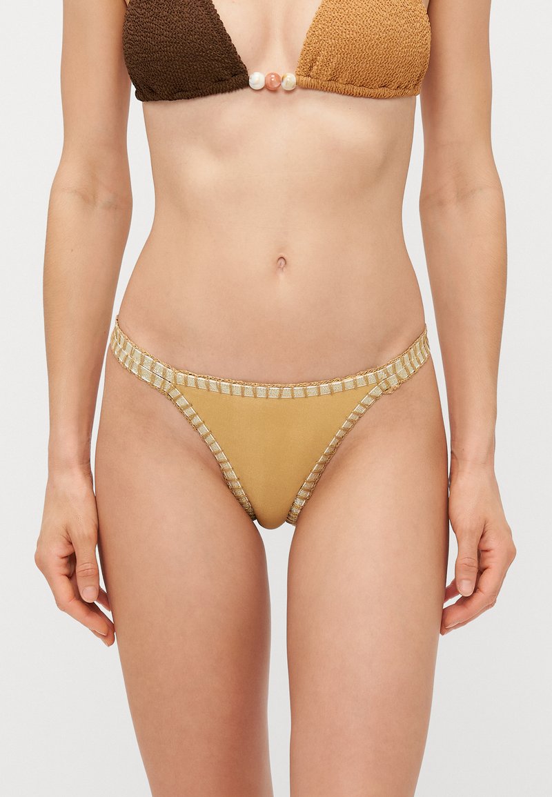 Kobieta ubrana w złote majtki bikini z metalicznym wykończeniem oraz dwukolorowy brązowy stanik bikini z ozdobnymi koralikami na środku.