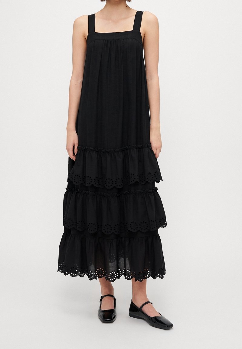 Femme portant une robe maxi noire sans manches à étages avec bordure en dentelle œillet et des ballerines noires Mary Jane, debout devant un fond uni.