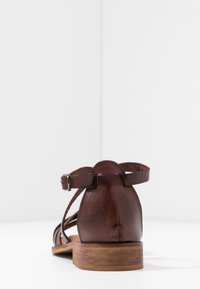 Sandale en cuir marron avec bride à la cheville et boucle argentée vue de dos sur une surface blanche avec un fond clair uni.