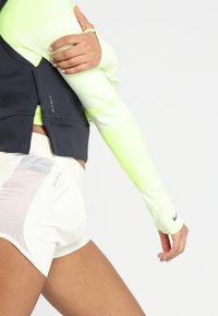 Zielona koszulka performance z długim rękawem i fakturowanymi rękawami, w zestawieniu z kontrastującymi białymi szortami z siateczkowymi akcentami i czystą kamizelką.