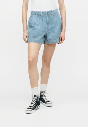 Persona con shorts a rayas azules y blancas, calcetines blancos, zapatillas altas negras y camiseta blanca, de pie contra un fondo liso.