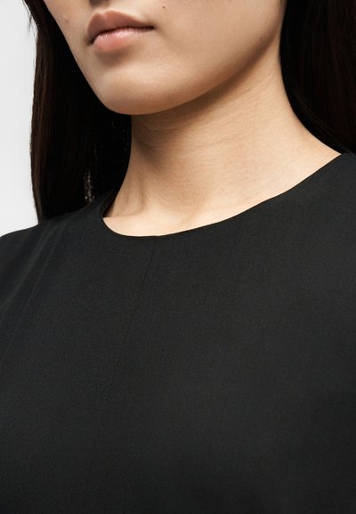 Inimese alaosa näo, kaela ja ülaosa rindkere lähivõte, kes kannab lihtsat musta ümmarguse kaelusega särki, pikkade tumedate juustega.