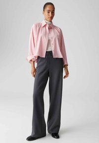 Roze, kort geknipte blouse met knopen en kraag, gecombineerd met losse, donkergrijze broek. Soepele textuur; broek heeft een tailleband met trekkoord.