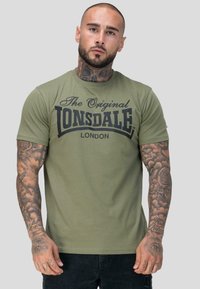 Habemega mees koos kaelatäitega, kandes oliivrohelist "The Original Lonsdale London" T-särki ja musti pükse, seismas ühetoonilise halli tausta ees.