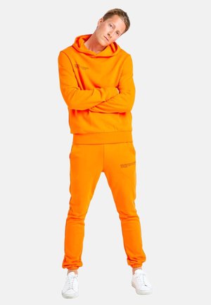 Jeune homme debout, bras croisés, portant un sweat à capuche orange vif et un jogging assorti avec des baskets blanches sur fond blanc.