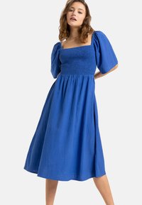 Vestido azul con escote cuadrado, cuerpo fruncido y mangas abullonadas. Hecho de una tela ligera, presenta una falda fluida que cae por debajo de la rodilla.