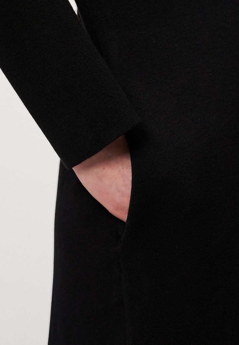Abito in maglia nera con maniche lunghe; presenta una tasca laterale e una texture liscia, con un focus sulla funzionalità e un design minimalista.