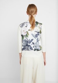 Žena se světle červenými vlasy ve spodním culíku má na sobě bílé dlouhé tričko s velkým květinovým potiskem na zadní straně a krémové široké kalhoty.