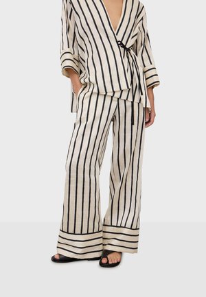 Kobieta ubrana w beżowe szerokie spodnie i dopasowany top kopertowy z pionowymi czarnymi paskami, zestawione z czarnymi sandałami na odkryte palce.