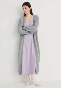 Cardigan lung gri peste o rochie lavandă, confecționată dintr-un material delicat. Modelul poartă ciorapi tricotati de culoare crem; designul este relaxat și stratificat.