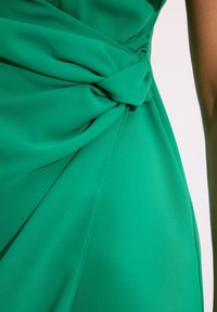 Zielona sukienka o gładkiej, miękkiej fakturze, z wyróżniającym się skręconym węzłem w talii oraz zwiewnym materiałem, który elegancko opada.
