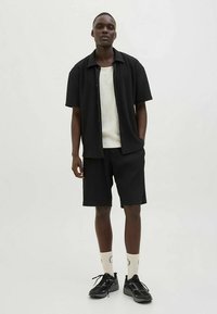 Cămașă neagră cu mâneci scurte și nasturi, cu un design texturat, combinată cu pantaloni scurți plisati negri și adidași negri. Maiou cu dungi, de culoare deschisă, pe dedesubt.