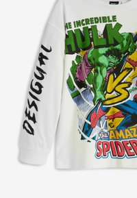 Baltos ilgomis rankovėmis marškiniai su komiksų stiliaus žaliu Hulk ir raudonu Spider-Man, kurie kovoja, tekstas "Nepaprastasis Hulk prieš Nuostabųjį Spider-Man" ir "DESIGUAL" ant rankovės.