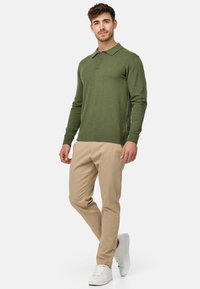 Suéter de color verde oliva con un cuello y dos botones, combinado con pantalones slim-fit en color beige y zapatillas blancas, de pie sobre un fondo liso.