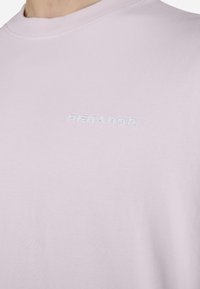T-shirt en coton rose clair avec un col rond, présentant une broderie blanche en relief du mot "Pecador" sur la poitrine. Texture lisse.