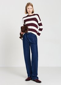 Junge Frau trägt einen burgunderfarbenen und weißen gestreiften Pullover, blaue Jeans, burgunderfarbene Stiefel und hält eine braune Jacke vor einem schlichten Hintergrund.