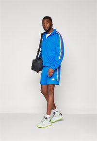 Tuta blu con giacca con zip e pantaloni corti, con bande multicolori. Sneakers chiare con accenti verdi e una borsa a tracolla nera.