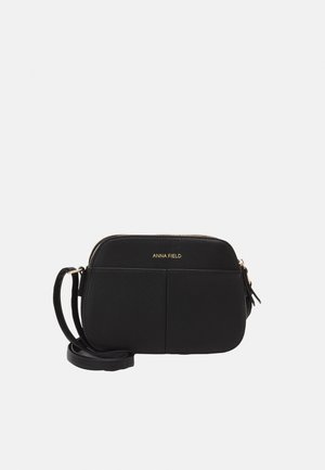 Sac bandoulière noir en matériau synthétique lisse, avec des bords arrondis, une fermeture éclair dorée et un logo de marque à l'avant.