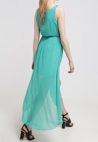 Akvamarínové šaty bez rukávů z lehkého, průhledného materiálu, stažené v pase, s dlouhým zadním lemem; doplněné černými vysokými podpatky.