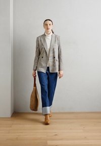 Prugasta blazer s reverom, bijeli turtleneck, široke plave traperice i bež čizme do gležnja; nosi pleteni, okrugli ručni kofer.