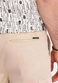 Chemise blanche à manches courtes avec un motif de texte noir, associée à un pantalon beige avec une poche arrière et une étiquette de marque.