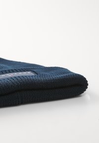 Navy blue knit beanie with a textured surface and rolled edge. Features a rectangular brand tag stitched onto the fabric.