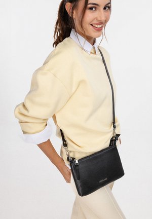 Lachende vrouw die een lichtgele trui en beige broek draagt, met een zwarte leren schoudertas met ritssluiting.