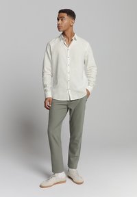 Licht beige overhemd met lange mouwen, gecombineerd met olijfgroene broek en beige sneakers, tegen een neutrale achtergrond.