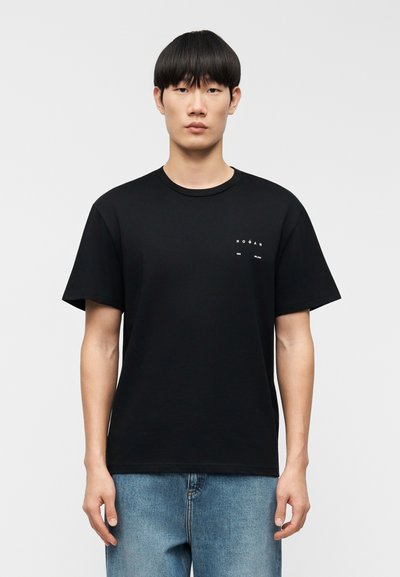 T-shirt en coton noir à manches courtes, col rond, avec un petit logo blanc imprimé sur le côté gauche de la poitrine. Porte avec un jean en denim bleu.