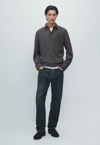 Grijs poloshirt met lange mouwen over een wit T-shirt, gecombineerd met donkerblauwe spijkerbroek en zwarte schoenen. Eenvoudig ontwerp, ontspannen pasvorm en gladde textuur.