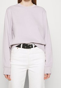 Mujer vistiendo una blusa de manga larga de color púrpura claro, metida en unos jeans blancos de tiro alto con un cinturón negro en la cintura.