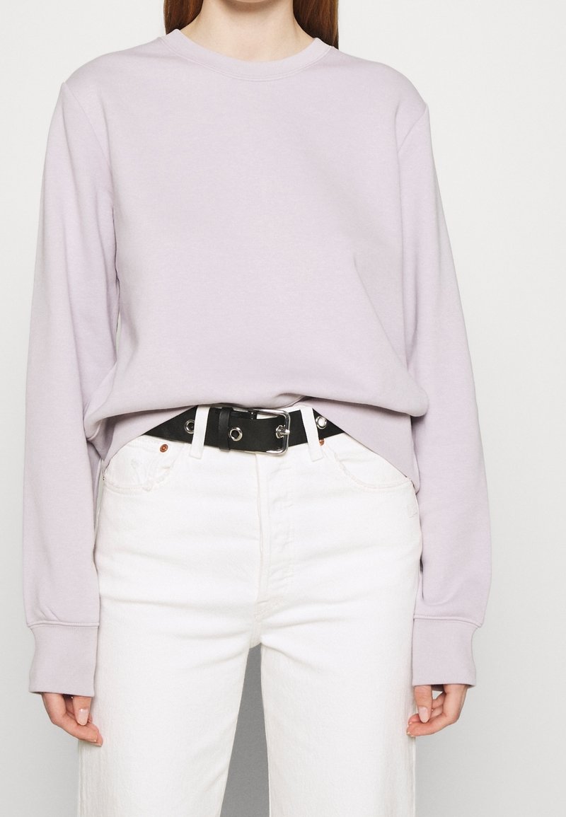 Mujer vistiendo una blusa de manga larga de color púrpura claro, metida en unos jeans blancos de tiro alto con un cinturón negro en la cintura.