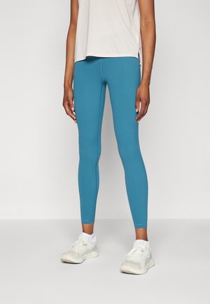 Blaue Leggings aus dehnbarem Material mit glatter Textur, kombiniert mit hellen Sportschuhen. Das Oberteil ist ein blasses, kurzärmeliges Shirt.