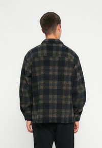 Jachetă cămașă cu carouri negre și maro, fabricată dintr-un material texturat, cu guler și mâneci lungi, având un croi lejer.