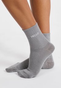 Calcetines grises de canalé con un ligero brillo, que presentan un texto en blanco que dice "HOLA". De longitud corta hasta el tobillo, textura suave y forma ajustada.