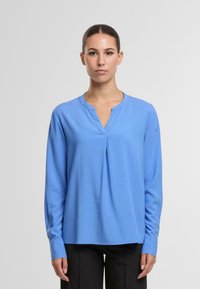 Mėlyna ilgomis rankovėmis bliusoniška su V formos iškirpte, pagaminta iš švelnios medžiagos. Priekyje yra raukšlių, pritaikyta silueto ir šiek tiek suapvalinta apačia.