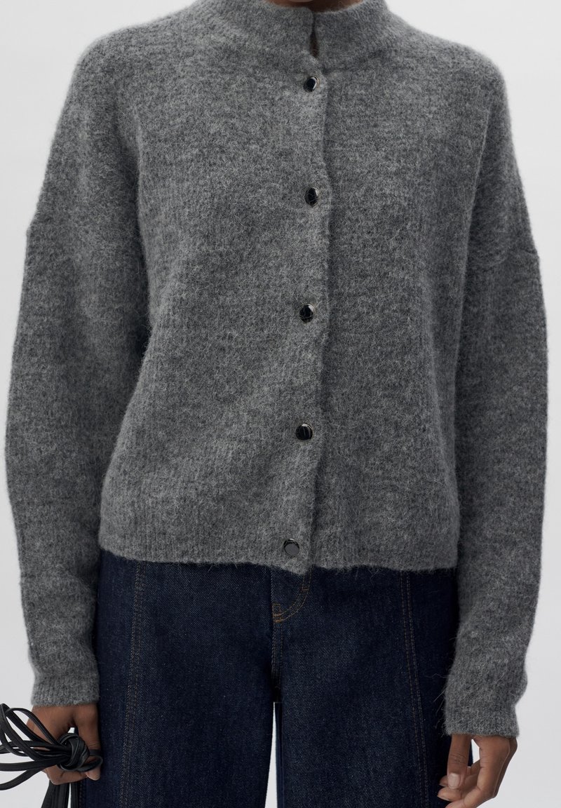 Personne portant un pull gris à boutons en tissu duveteux avec manches longues et un jean bleu foncé, tenant un câble noir enroulé dans une main.