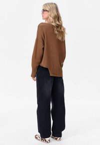 Bruine gebreide trui met een losse pasvorm, geribbelde zoom, gecombineerd met donkere wijde spijkerbroeken en gevlekte instapschoenen. Model poseert van achteren.