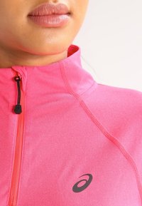 Nahaufnahme des unteren Gesichts und des Halses einer Person, die ein hellrosa Reißverschluss-Sportoberteil mit einem Asics-Logo auf der Brust trägt.