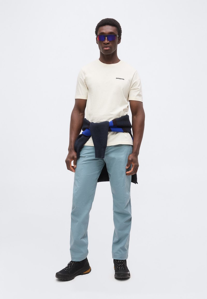 Mann mit Sonnenbrille, beigem Patagonia T-Shirt, hellblauer Hose, schwarzen Wanderschuhen und einem dunkelblauen Pullover um die Taille gebunden, stehend vor weißem Hintergrund.