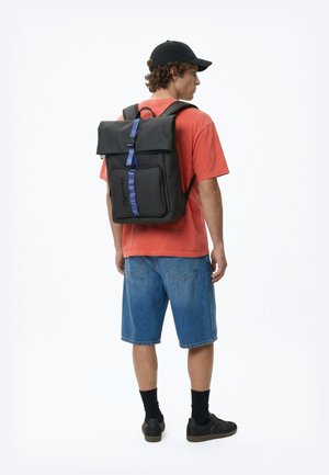 Nuori mies, jolla on musta lippis, oranssi t-paita, siniset farkkushortsit ja mustat tennarit, seisoo oikealle päin katsellen mustan repun kanssa, jossa on sininen hihna.