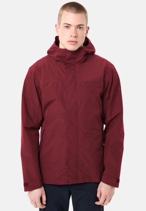 Jeune homme portant une veste à capuche bordeaux zippée, à manches longues et avec des poches, debout devant un fond clair uni.