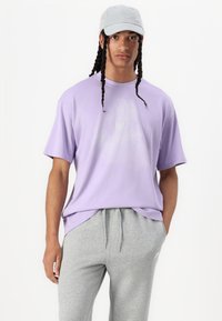 Φαρδιά μωβ κοντομάνικη μπλούζα με διακριτικό λευκό μοτίβο και μικρό κίτρινο λογότυπο· συνδυάζεται με γκρι παντελόνι φόρμας. Ύφασμα από βαμβακερό μείγμα.