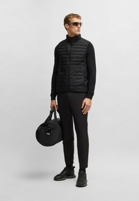 Schwarze gesteppte Weste mit hohem Kragen, getragen über einem schwarzen Langarmoberteil, kombiniert mit schwarzen Hosen und Turnschuhen; hält eine runde schwarze Tasche.