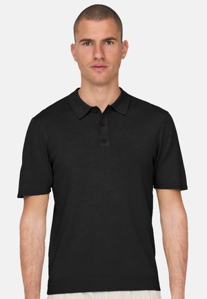 Lyhytukkainen mies, jolla on musta lyhythihainen poolopaita, jossa on kolme nappia, seisomassa yksinkertaisen vaalean taustan edessä.