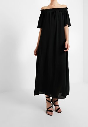 Vrouw die een lange zwarte off-shoulder jurk draagt en zwarte hoge hakken met gekruiste bandjes, staand tegen een effen achtergrond.