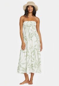 Kvinne iført ermeløs hvit kjole med grønn blad mønster og beige vevd bøttehatt, som står barbeint mot hvit bakgrunn.