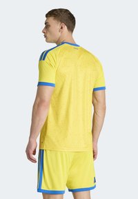 Maglia da atletica gialla con accenti blu, caratterizzata da un motivo geometrico texturizzato sul retro. Indossata con pantaloncini gialli abbinati.
